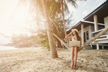 Woman sitting in hammock working with laptop computer on tropical island beach under palm tree
