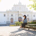 Young Hispanic woman sitting watching her phone in front of the San Jose cathedral in Antigua Guatemala-tourist traveling in colonial city-woman in the central park of Antigua Guatemala