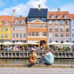 Two people sit by the canal in Copenhagen, enjoying the sunshine and the view of colorful buildings. Copenhagen Denmark