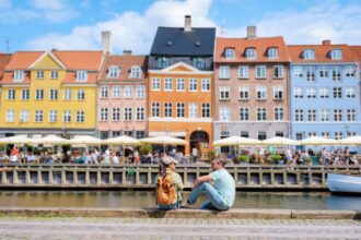 Two people sit by the canal in Copenhagen, enjoying the sunshine and the view of colorful buildings. Copenhagen Denmark