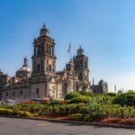 The Metropolitan Cathedral in Mexico City, surrounded by colorful gardens and the Mexican flag, under a bright clear blue sky in Zocalo Square.