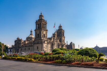 The Metropolitan Cathedral in Mexico City, surrounded by colorful gardens and the Mexican flag, under a bright clear blue sky in Zocalo Square.